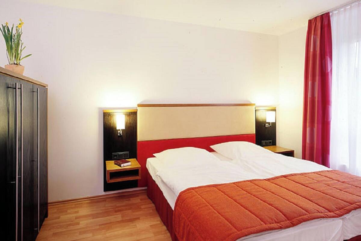 Une chambre d’hôtel avec un lit, un plancher en bois, des tables de chevet, une lampe, une armoire et une fenêtre avec des rideaux.