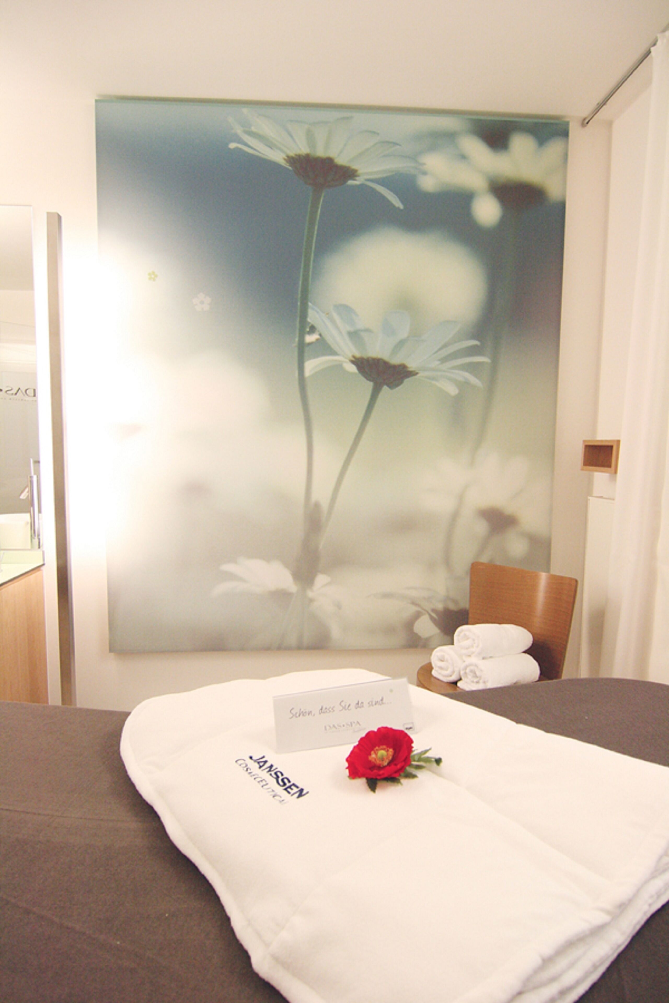 Une chambre d’hôtel avec un lit, un tableau représentant des fleurs accroché au mur, un porte-nom et une rose rouge posée sur le lit.