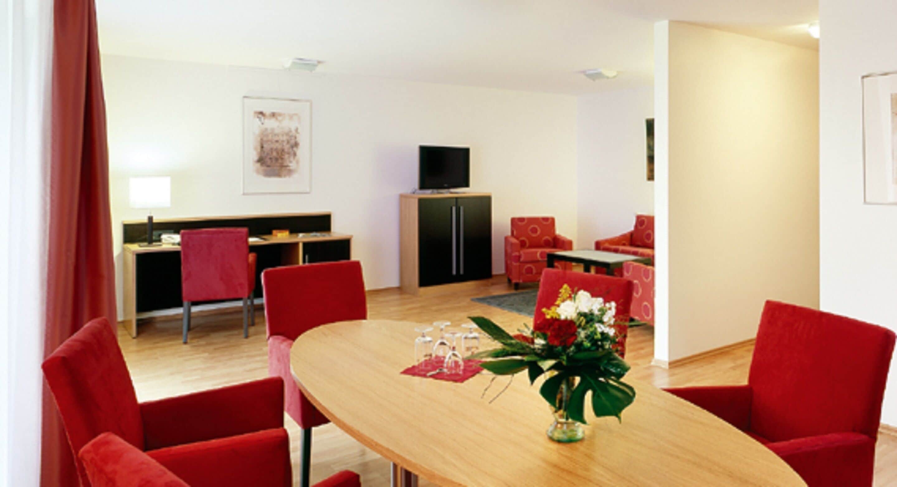 A modern living room with a wooden dining table, red chairs, a flat-screen TV, and a small kitchen area.