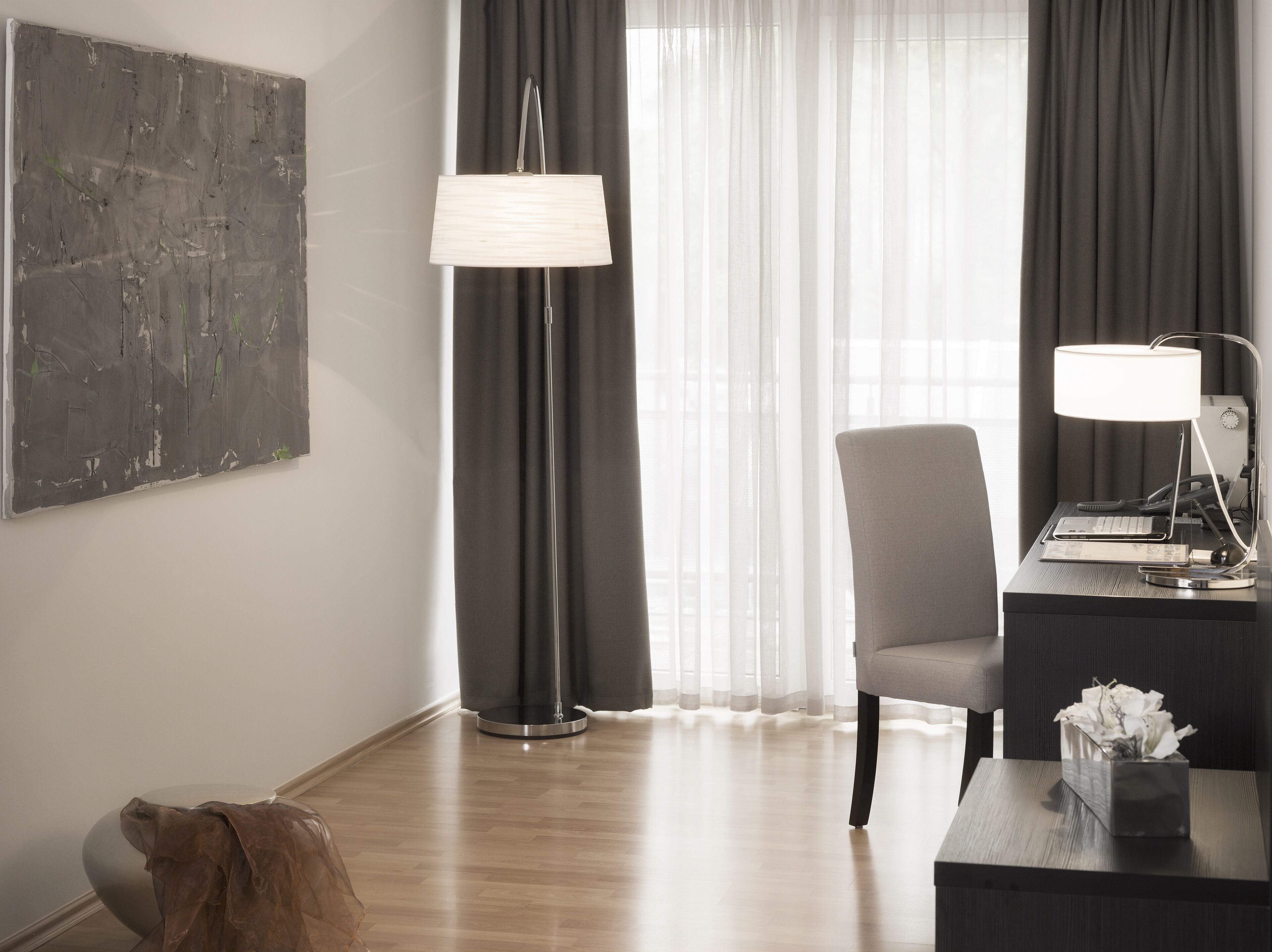 Quarto de hotel moderno com uma grande pintura abstrata, uma luminária de pé, uma escrivaninha com luminária, uma cadeira e uma pequena mesa com caixa de lenços.