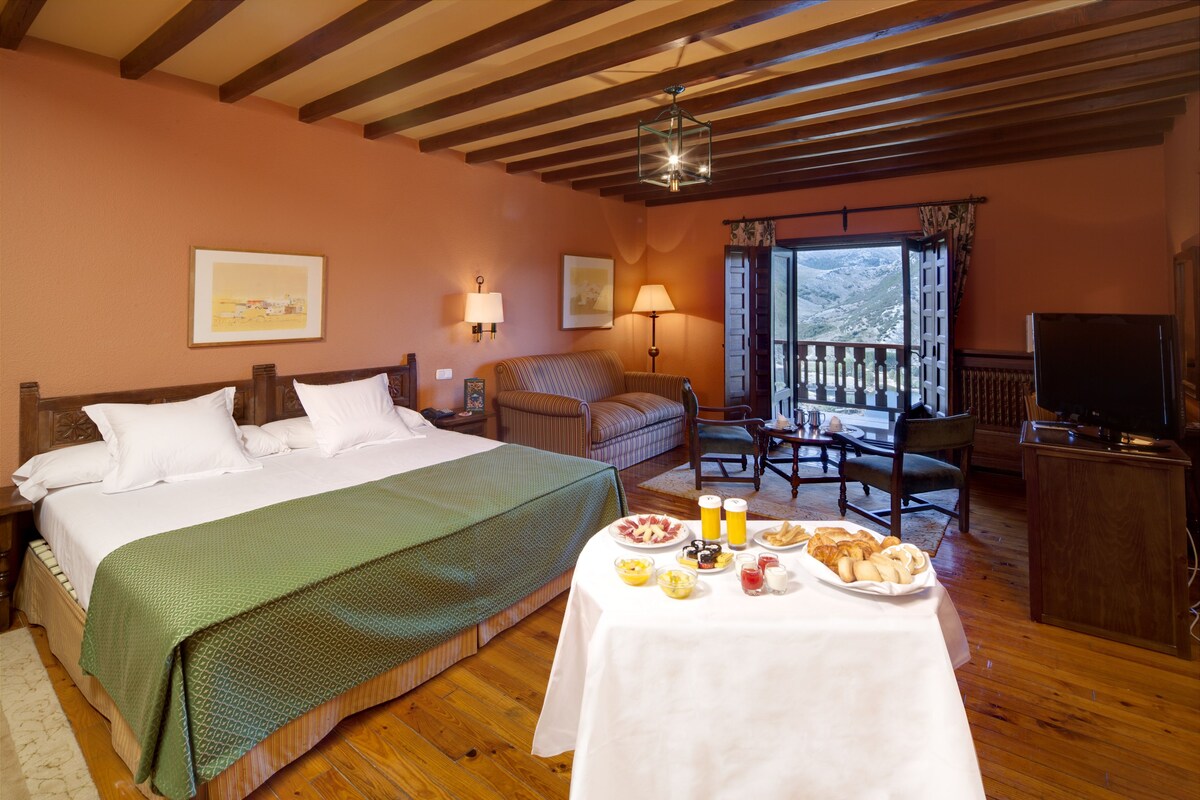 A hotel room with a large bed, a dining table with breakfast, a balcony with a view, and a TV.