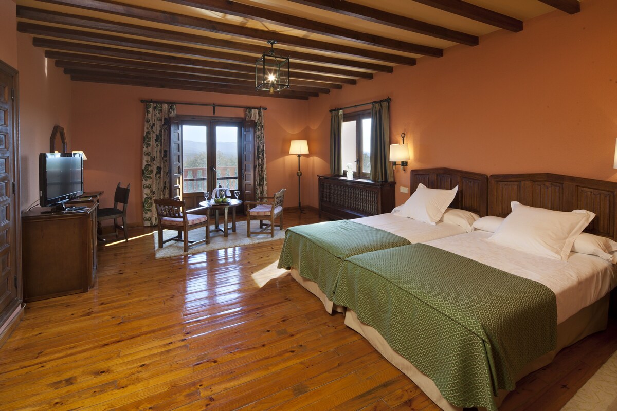 A hotel room with a wooden floor, a bed with two pillows, a desk with a television, and a window with curtains.