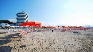 A beach with rows of orange umbrellas and white lounge chairs. A tall hotel building is visible in the background.