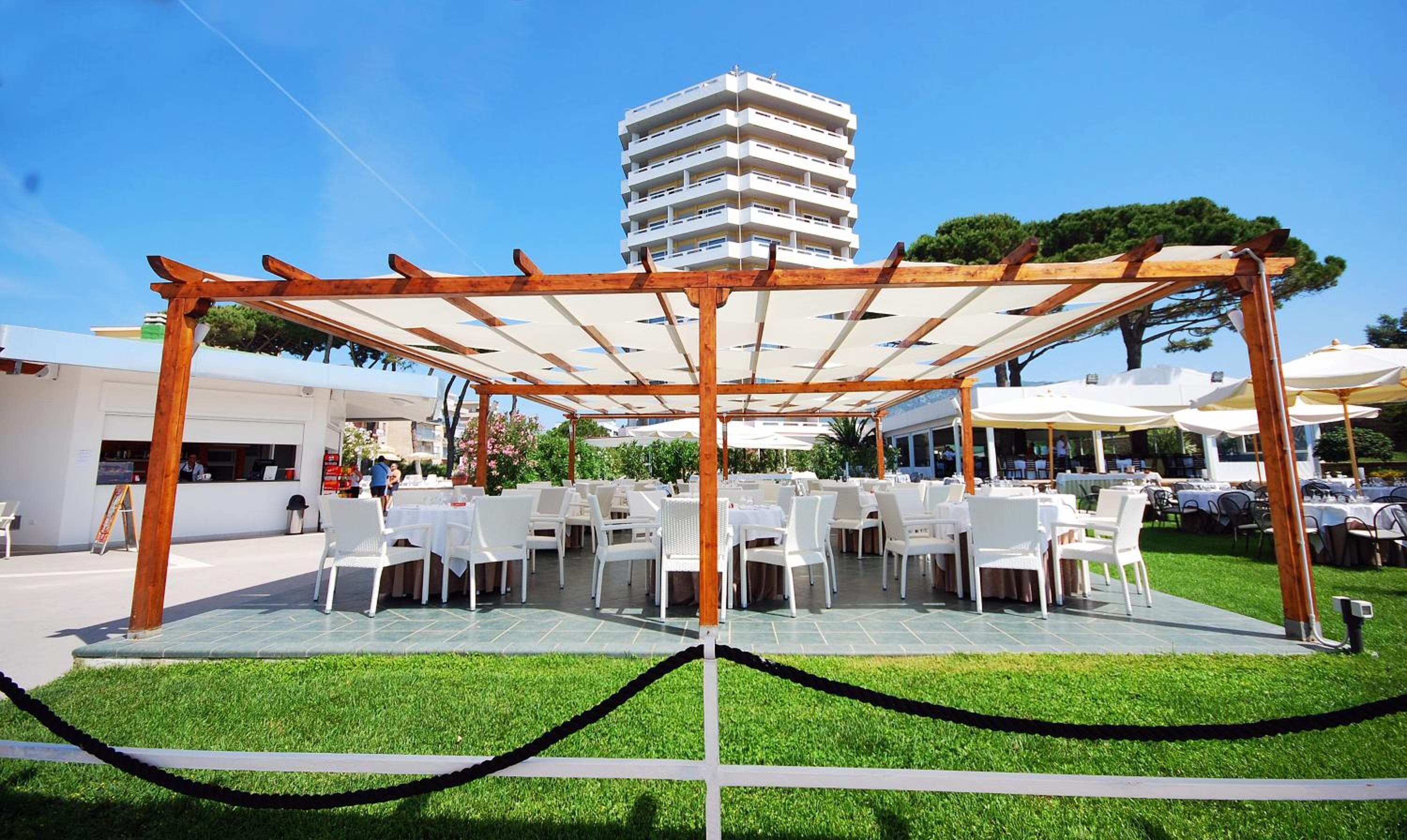 Un espace de restauration en plein air avec des tables et des chaises blanches, une pergola en bois et une vue sur un grand immeuble en arrière-plan.