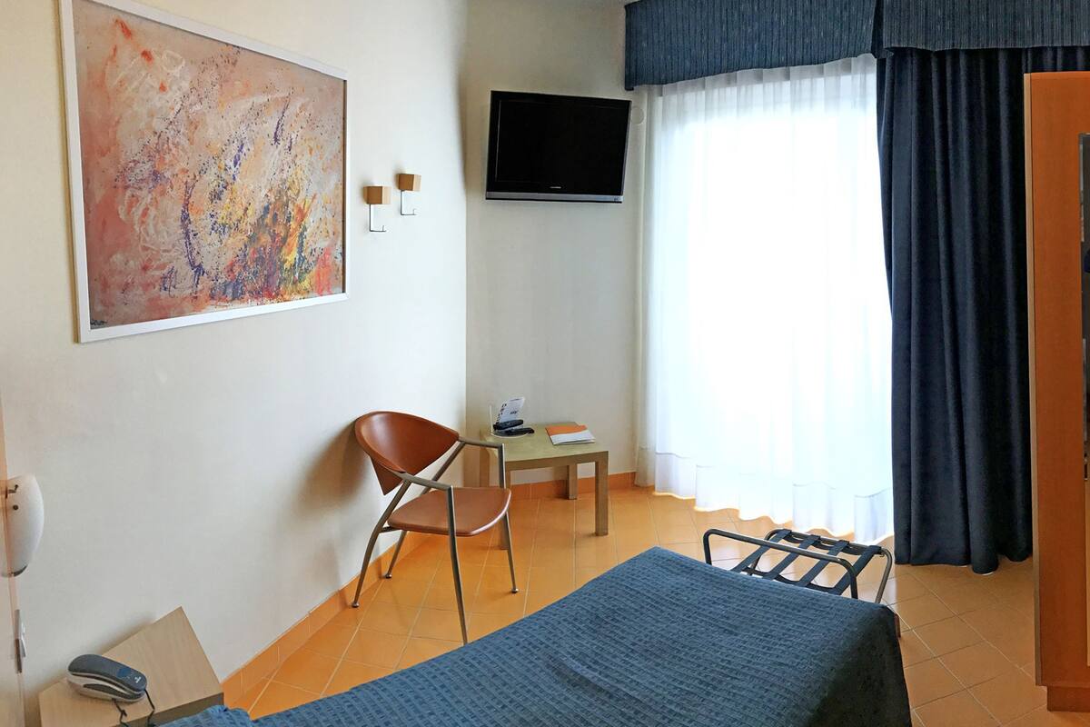 A hotel room with a bed, a desk, a chair, a TV, and a painting on the wall.
