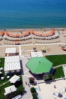 Vista aérea de un complejo turístico frente a la playa, con zona de piscina, mobiliario exterior y sombrillas.