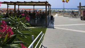 Ett kafé vid stranden med uteservering, en träpergola och utsikt över havet.