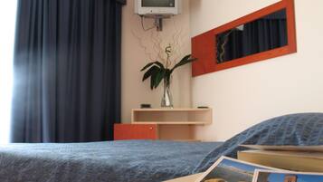 Habitación de hotel con cama, televisor montado en la pared, una planta y una mesita con una postal.