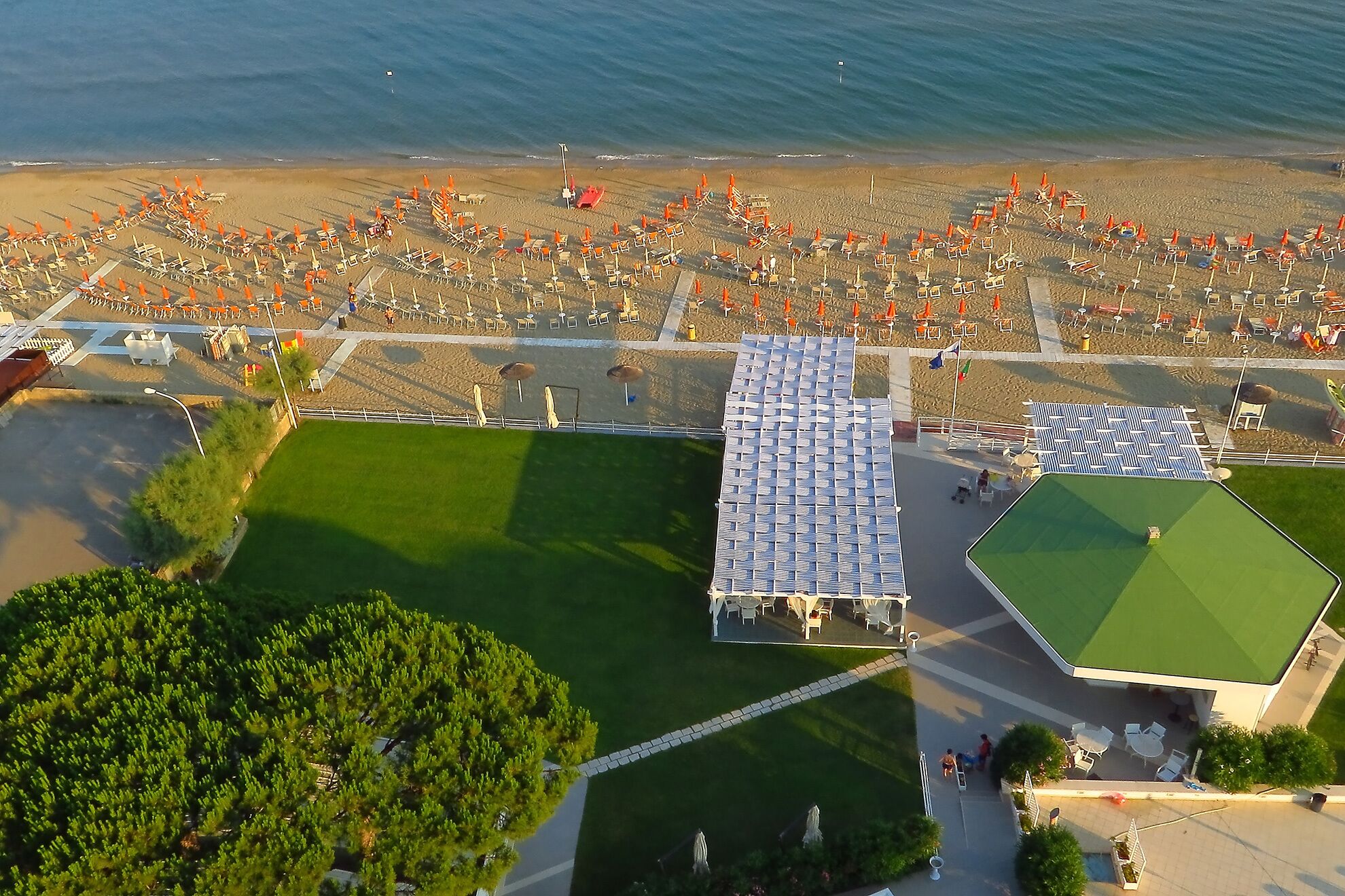 Vista aérea de una playa con filas de sombrillas y tumbonas, un área verde con césped y un edificio con techo verde.