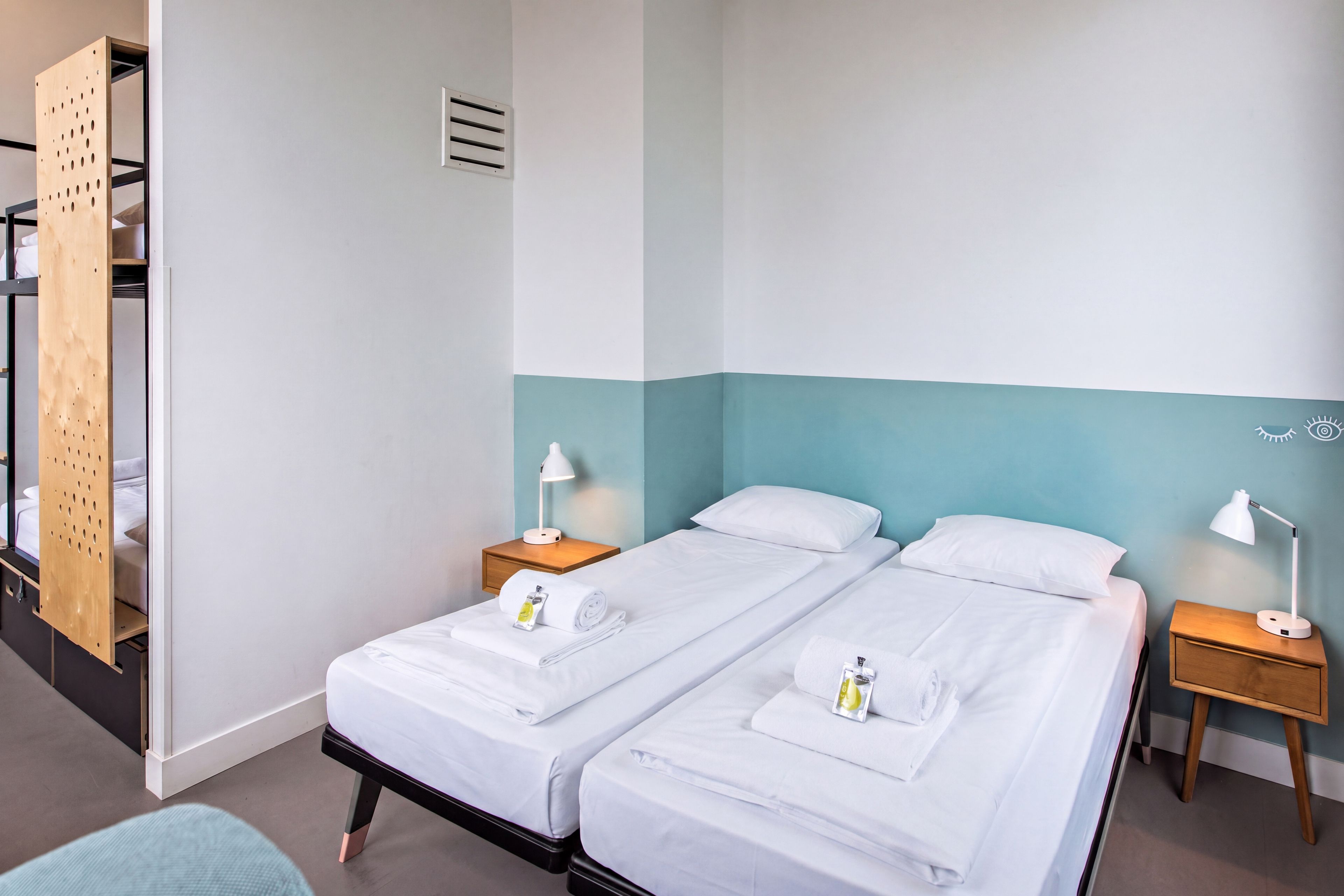 A hotel room with two beds, a wooden wardrobe, a nightstand with a lamp, and a wall-mounted vent.