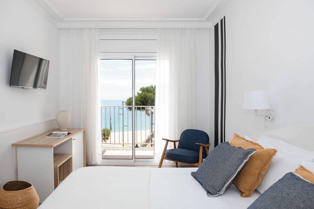 A hotel room with a bed, a desk, a chair, a TV, and a balcony with a view of the sea.