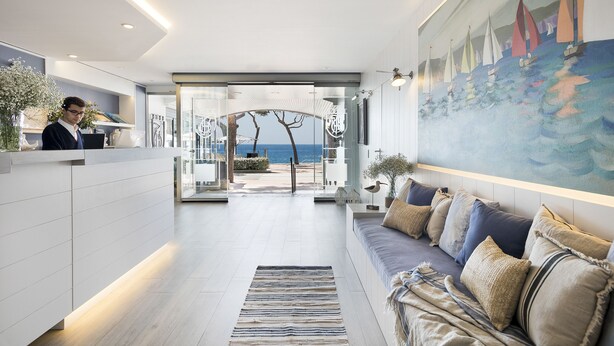 Un hall d’hôtel moderne avec un comptoir d’accueil, un canapé avec des coussins, un tableau représentant des voiliers et une vue sur la mer à travers les portes vitrées.
