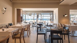 Un restaurant où les tables sont dressées pour le service, de grandes fenêtres offrant une vue sur la mer et un décor intérieur moderne.