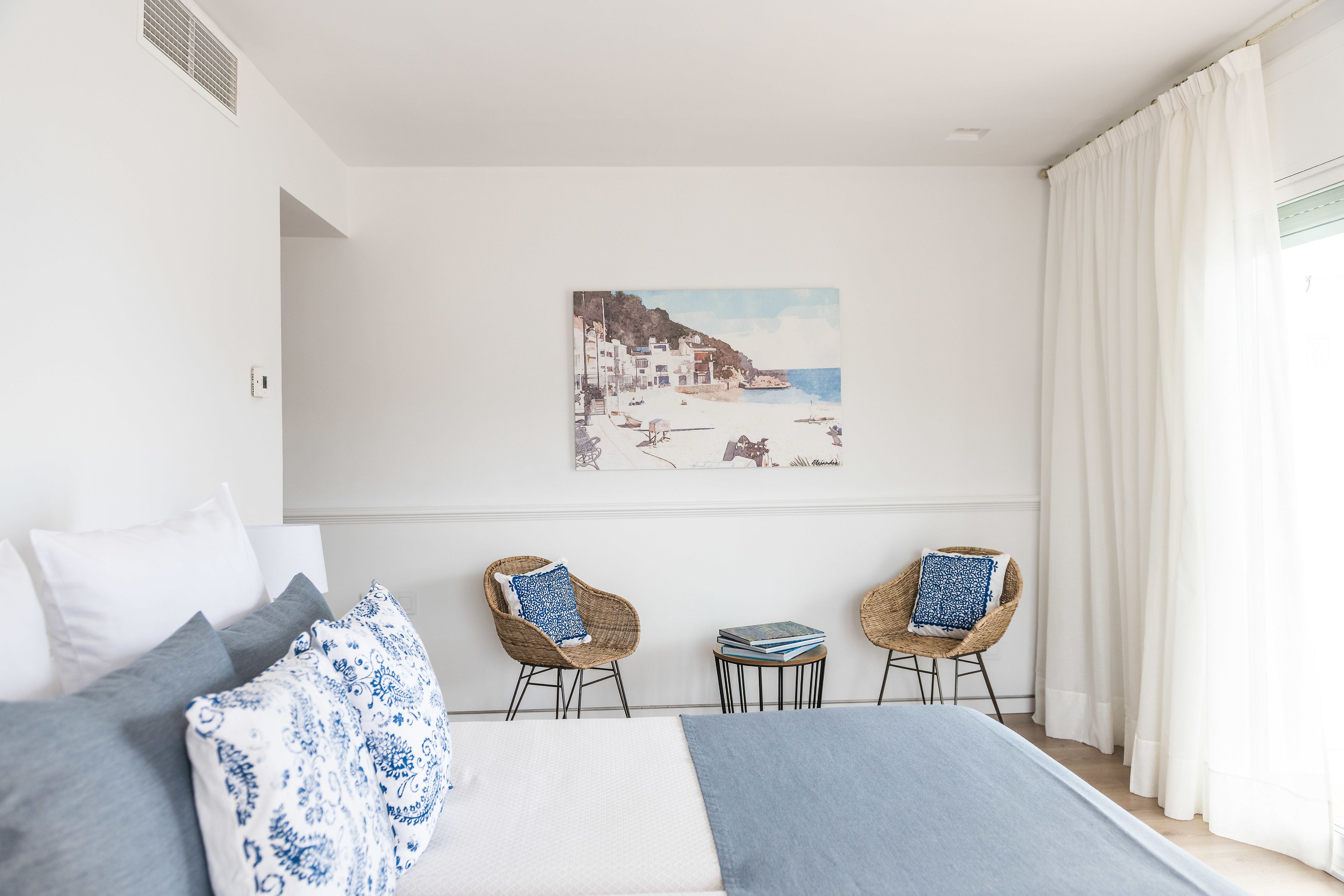 A bedroom with a bed, two wicker chairs, a small table, and a painting of a coastal scene.