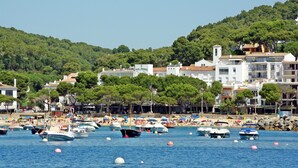 Une ville côtière où de nombreux bateaux sont amarrés le long du port, une plage animée avec des gens et des parasols, et des bâtiments dotés de balcons donnant sur la mer.