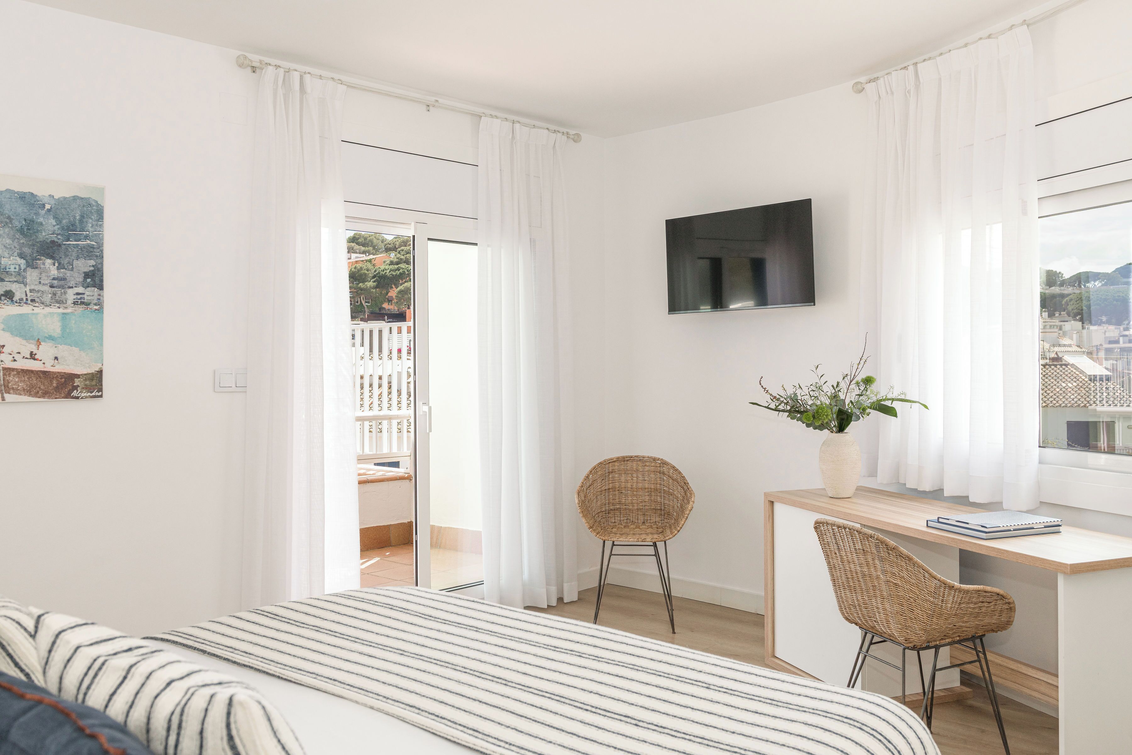 A modern bedroom with a bed, a desk, two wicker chairs, a TV, and a view of a balcony with a pool.