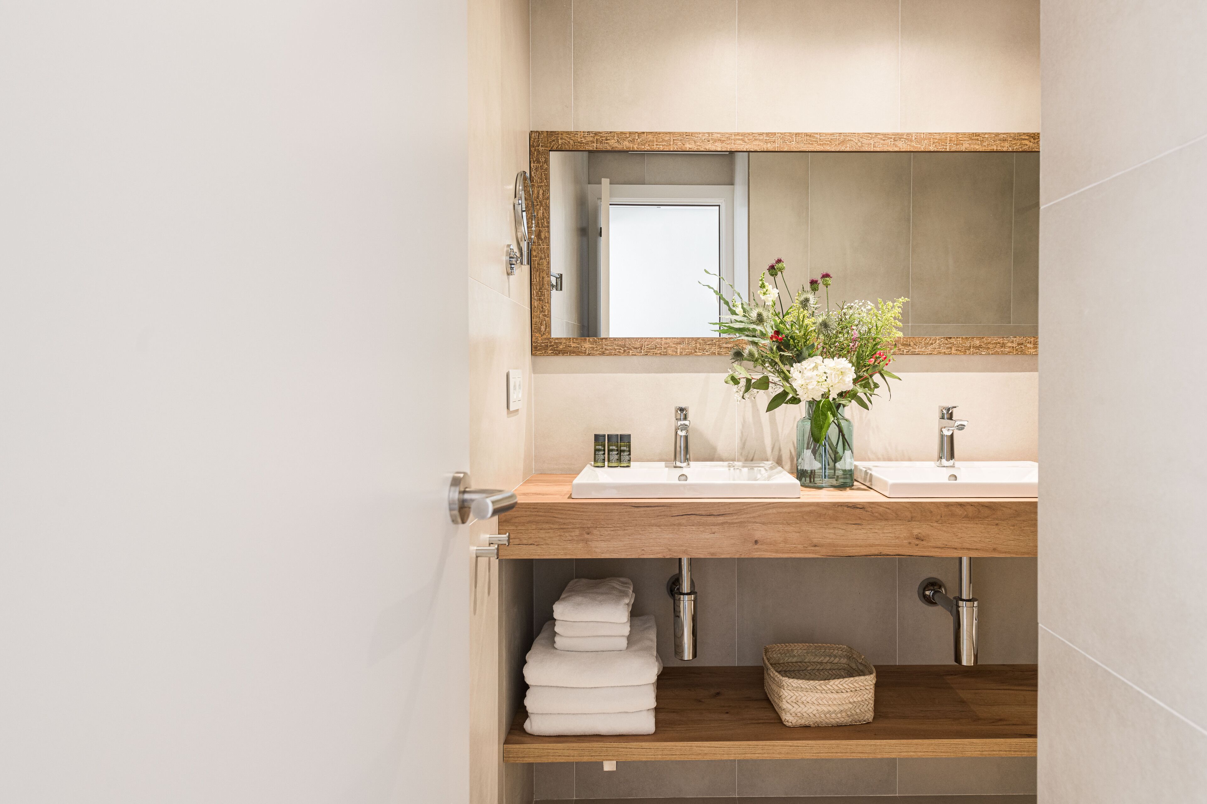 Un baño moderno con tocador de madera, dos lavamanos, un espejo grande y un florero con flores.