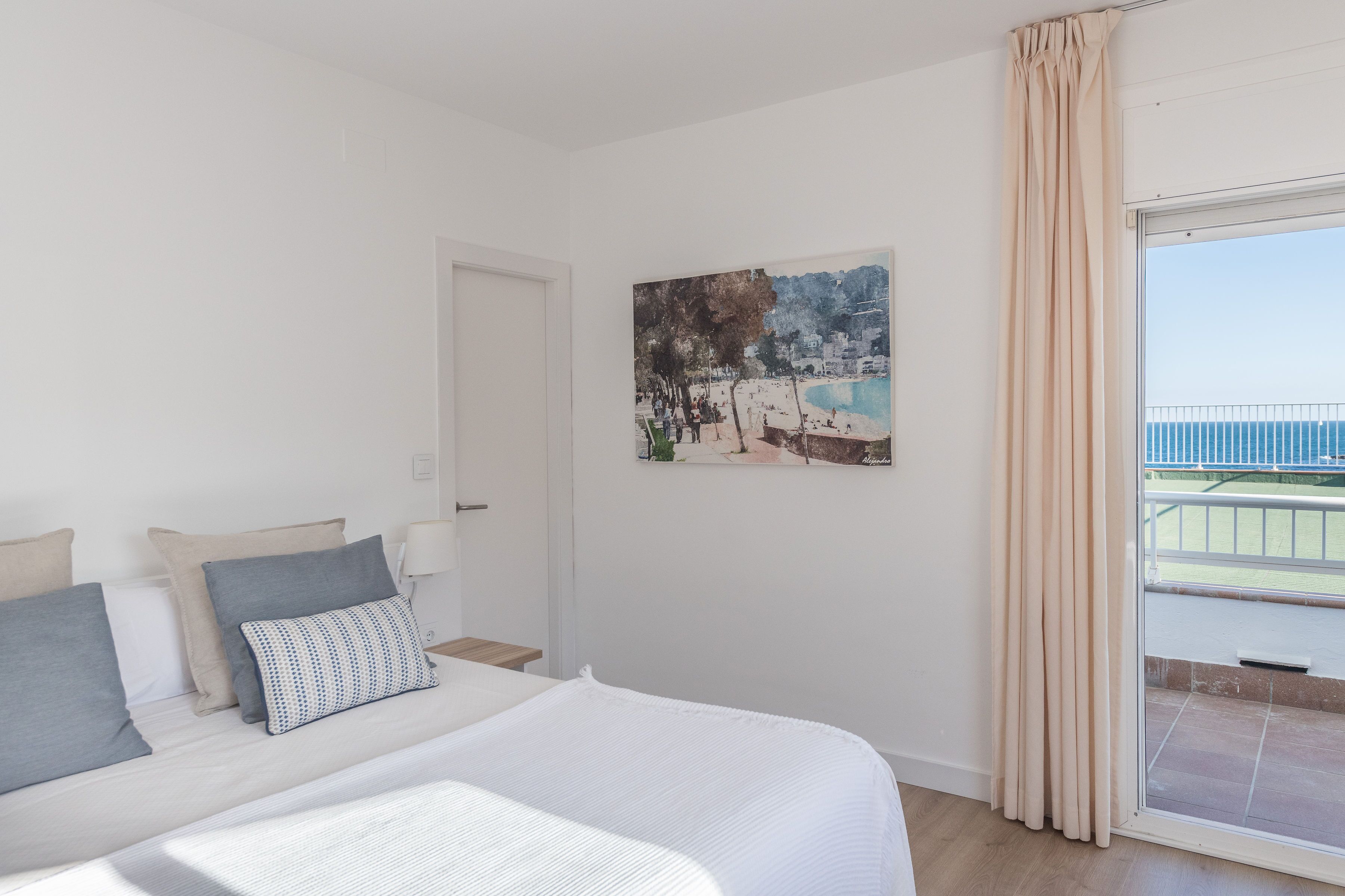 Een hotelkamer met een bed, een schilderij van een strand en een raam met gordijnen, met uitzicht op zee.