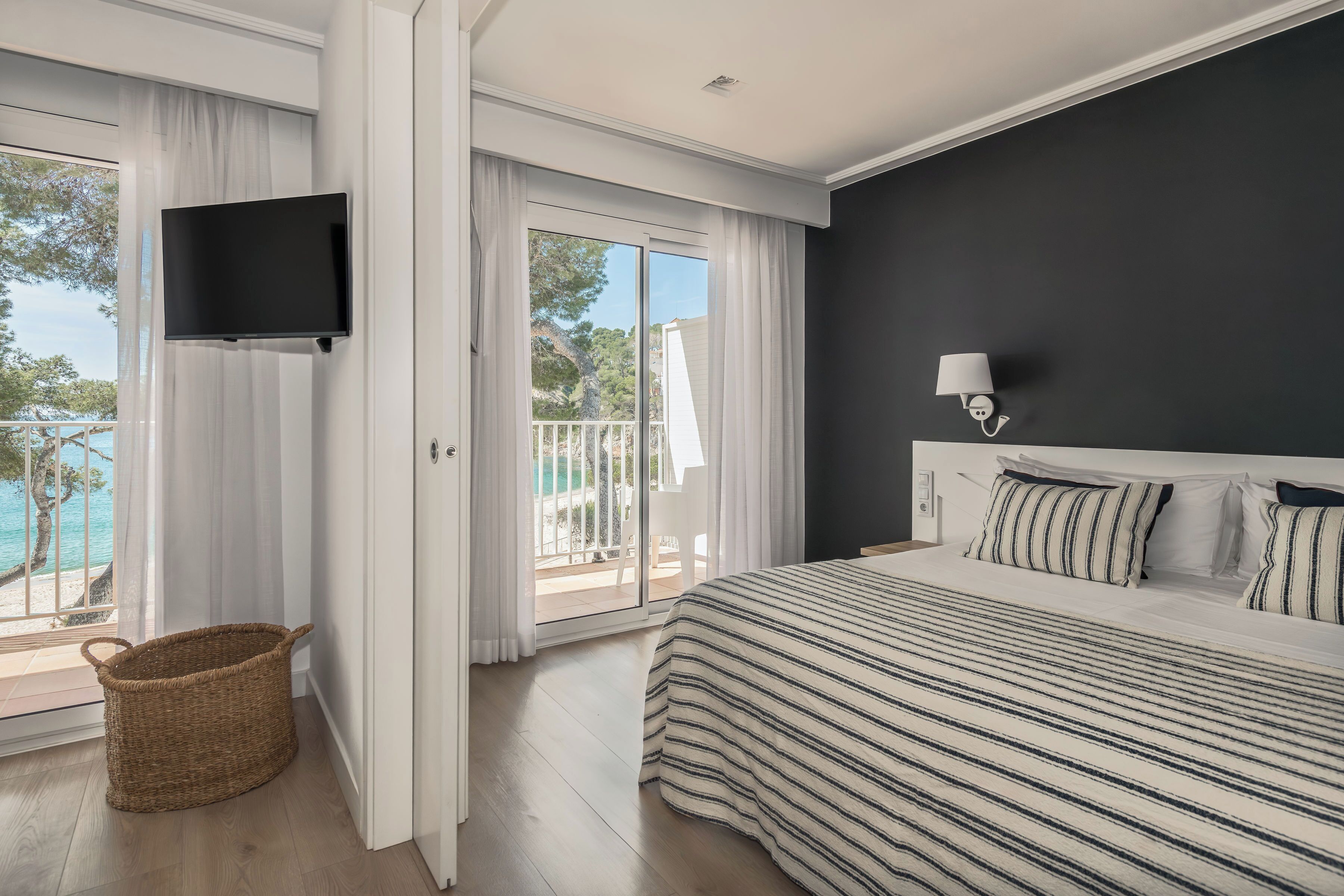 A bedroom with a bed, a TV mounted on the wall, a balcony with a view of the sea, and a wicker basket on the floor.