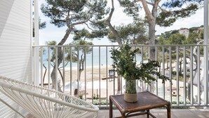 Un balcon avec une chaise en osier blanche, une table en bois ornée d’un pot de fleurs, et une vue sur la plage et les arbres.