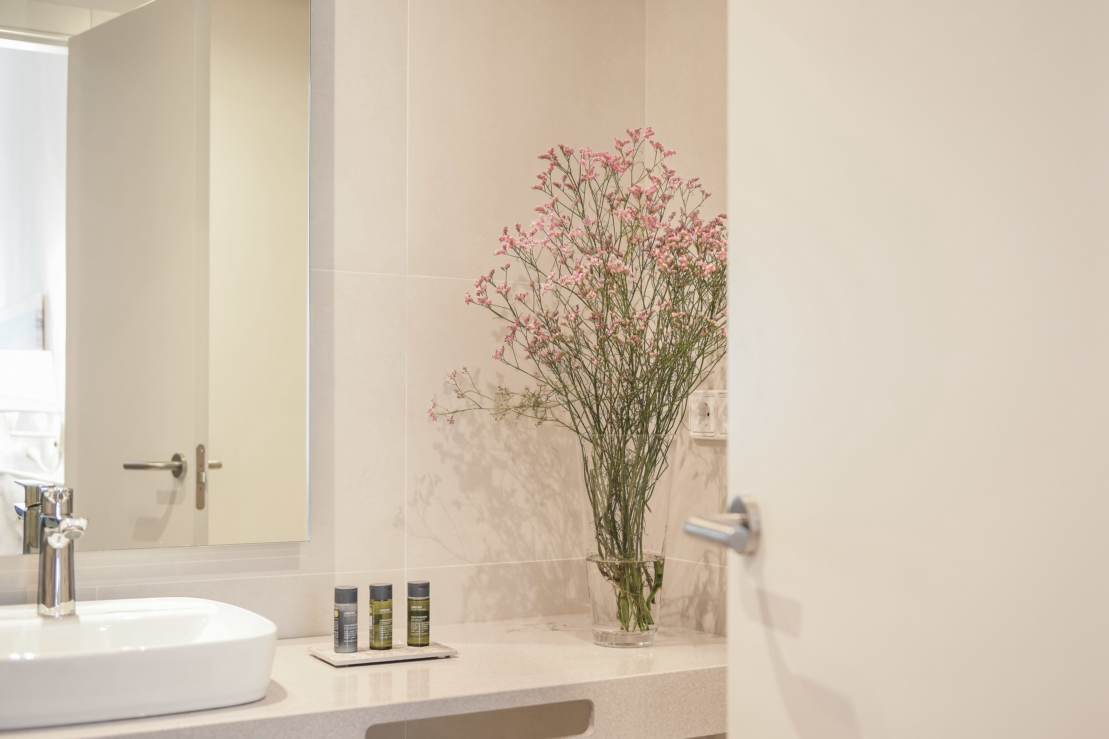 Une salle de bain avec un lavabo blanc, un miroir et un vase contenant des fleurs roses, posé sur le comptoir.