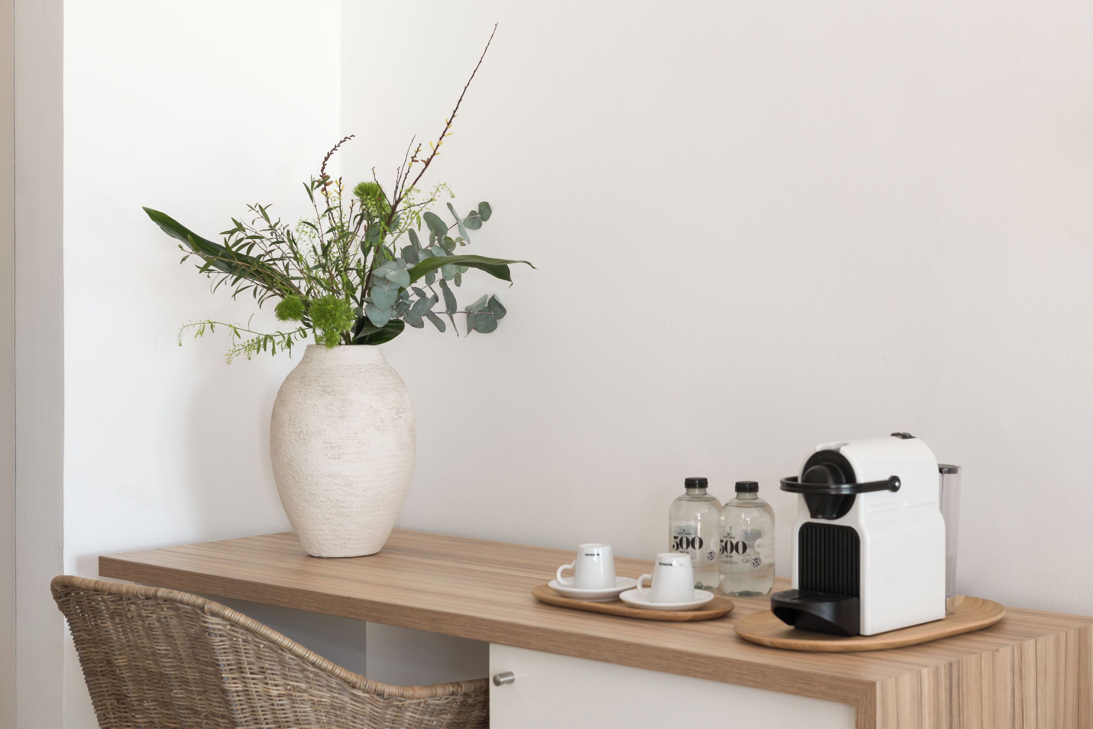 Un bureau en bois sur lequel se trouve un vase blanc contenant des plantes vertes, deux tasses blanches et une machine à café.