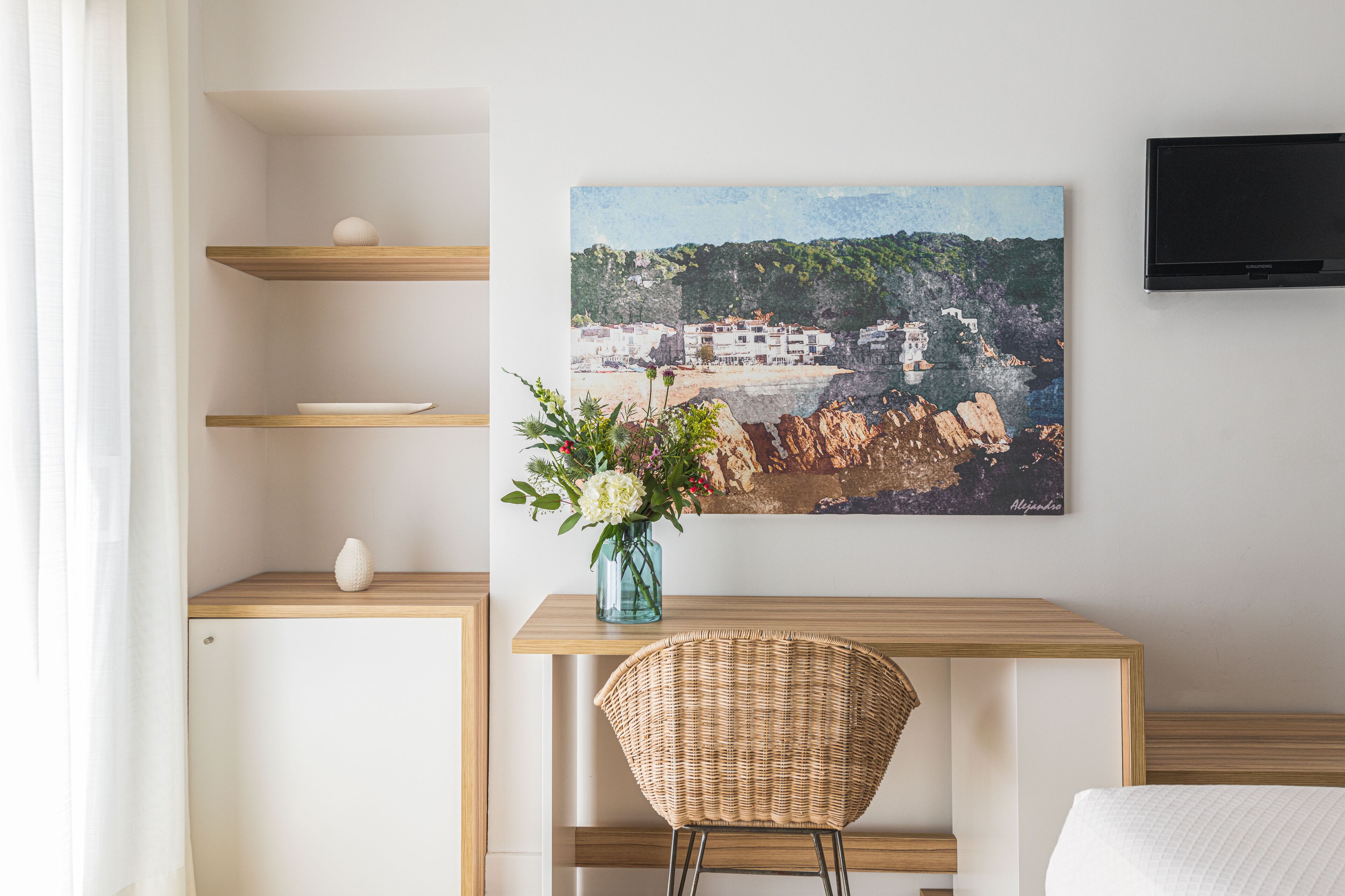 Ett hotellrum med ett skrivbord i trä, en fåtölj i rotting, en vit säng, en tv som är monterad på väggen och en tavla som föreställer en kuststad.