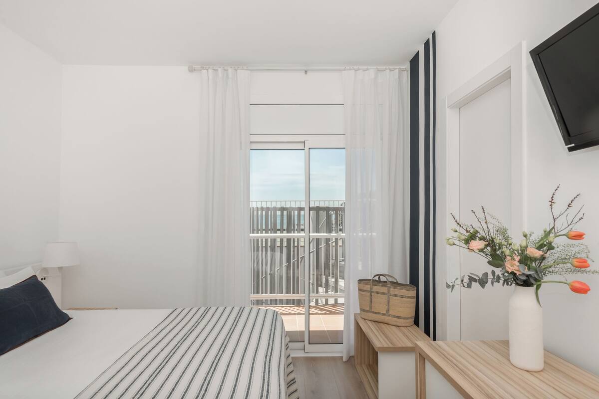 A bedroom with a bed, a nightstand, a TV mounted on the wall, a window with curtains, and a balcony with a railing.