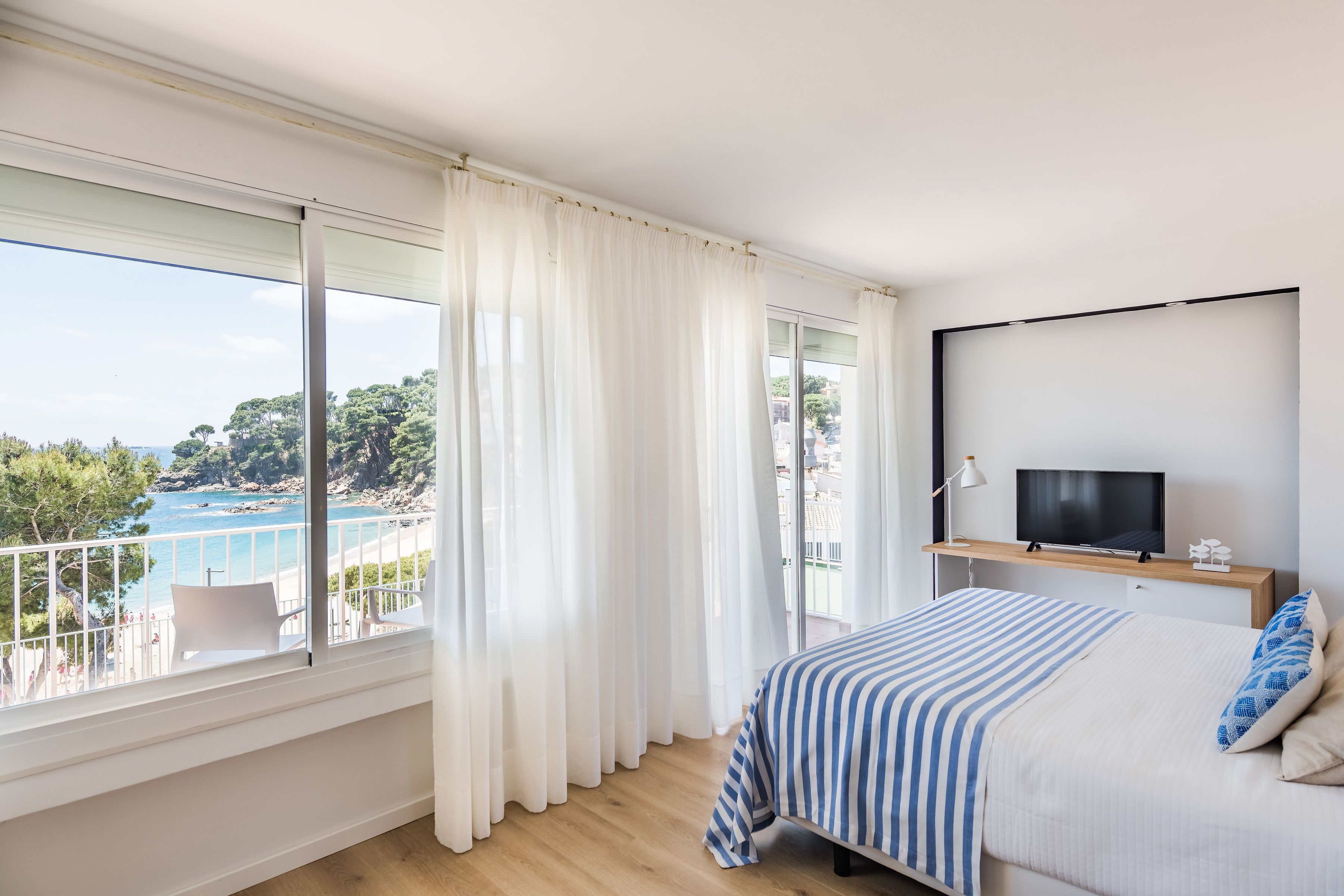 A bedroom with a large window, a bed, a TV on a wooden shelf, and a balcony with a chair.
