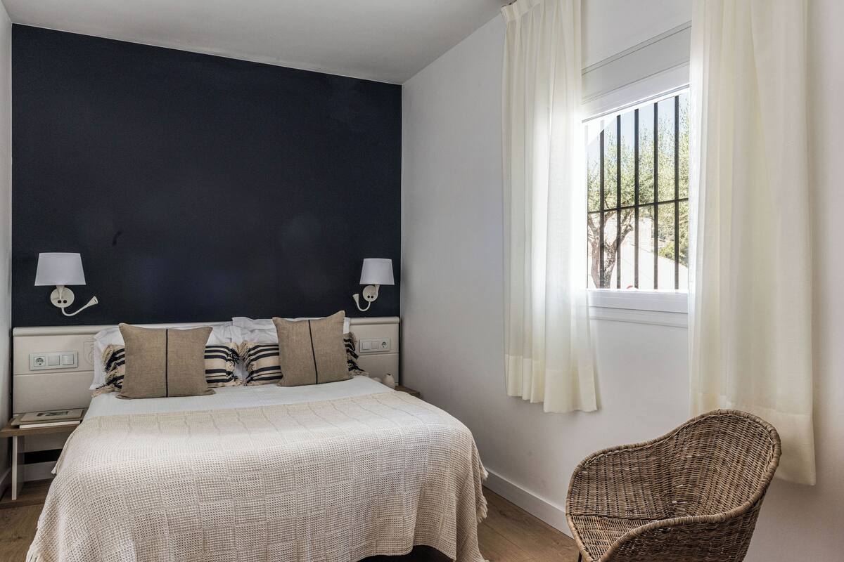 A bedroom with a bed, two bedside lamps, a wicker chair, a window with curtains, and a dark accent wall.