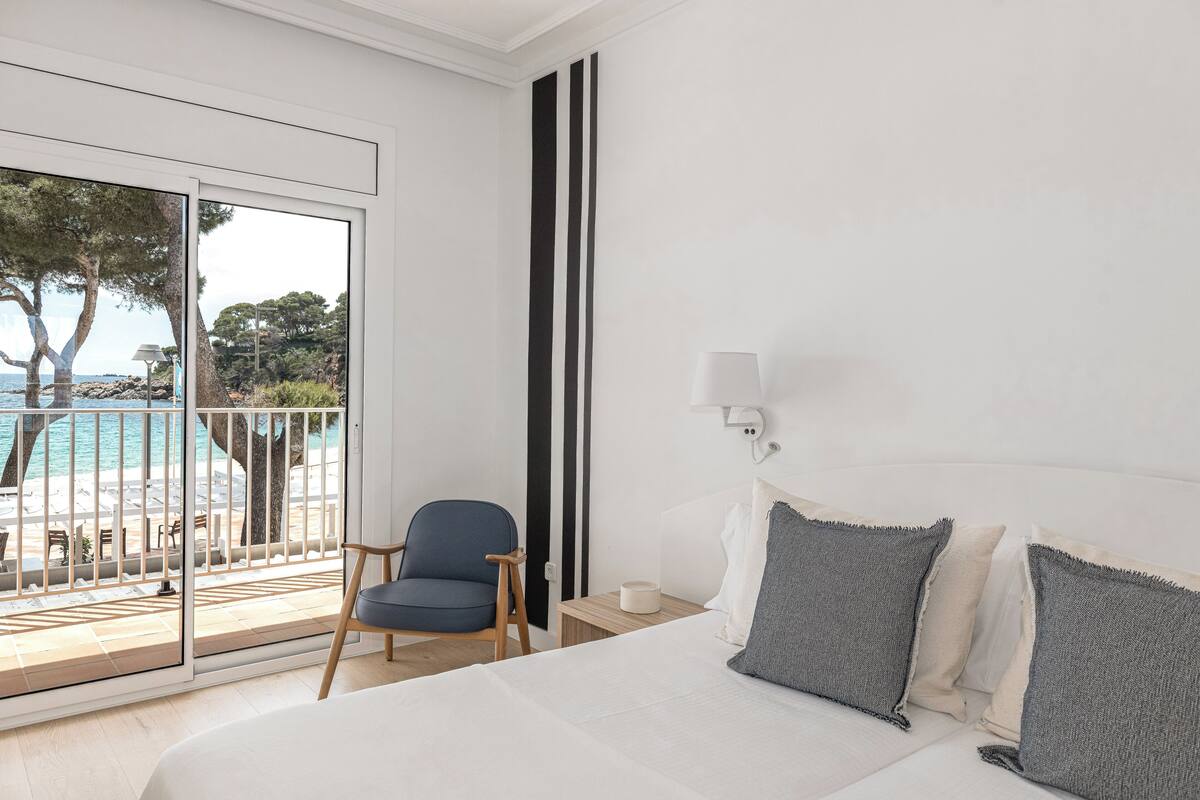 A hotel room with a bed, a chair, a nightstand, a lamp, and a view of a pool and trees.