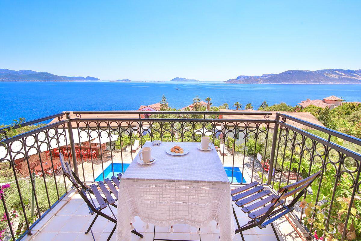 A balcony with a table set for two, overlooking a scenic view of the sea and distant hills.