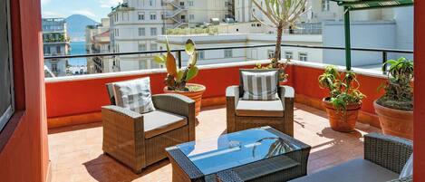 A balcony with wicker furniture, potted plants, and a view of buildings and a mountain.