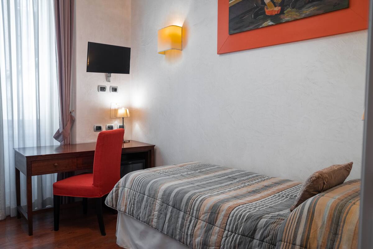 A hotel room with a wooden desk, a red chair, a bed with striped bedding, a mounted TV, a wall-mounted lamp, and a painting on the wall.