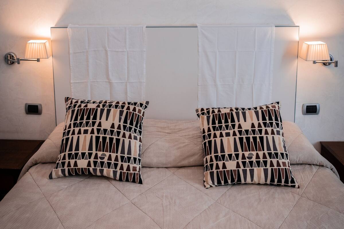 A bedroom with a bed, two patterned pillows, a painting above the bed, and wall-mounted lamps.