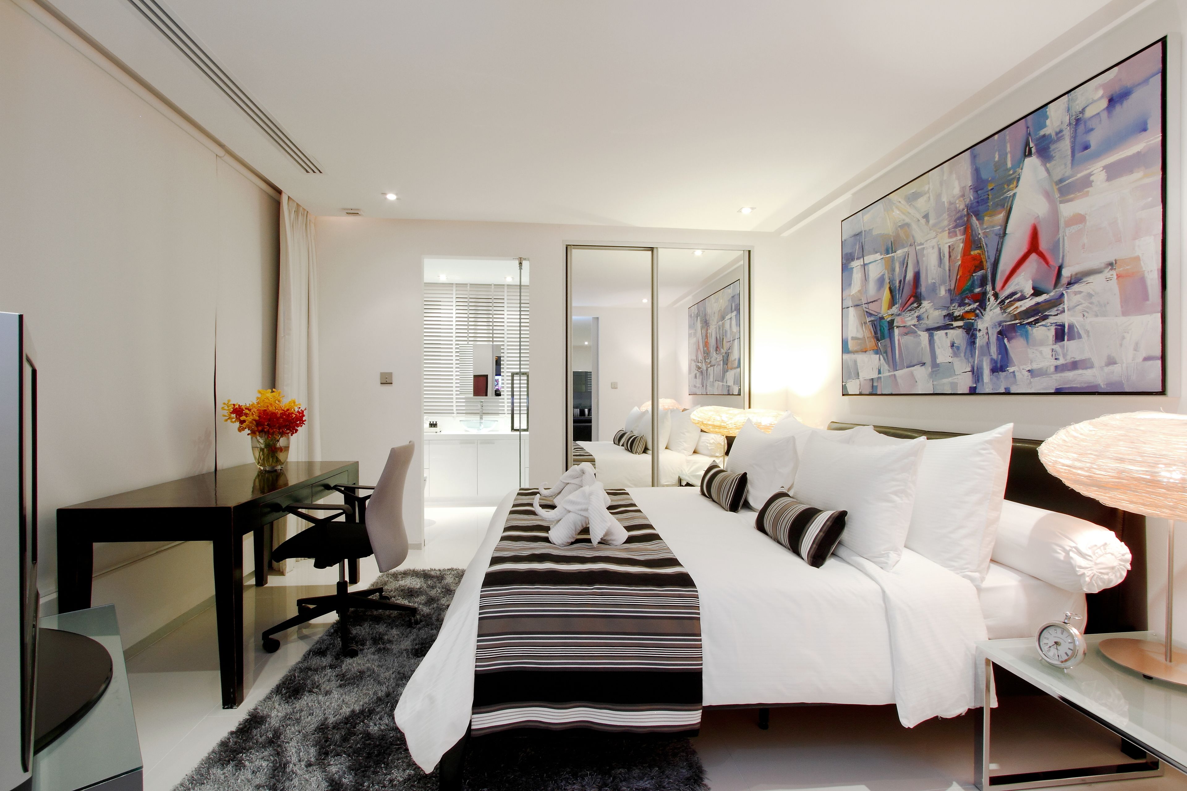 A modern hotel room with a large bed, a desk, a chair, a TV, and a painting on the wall.