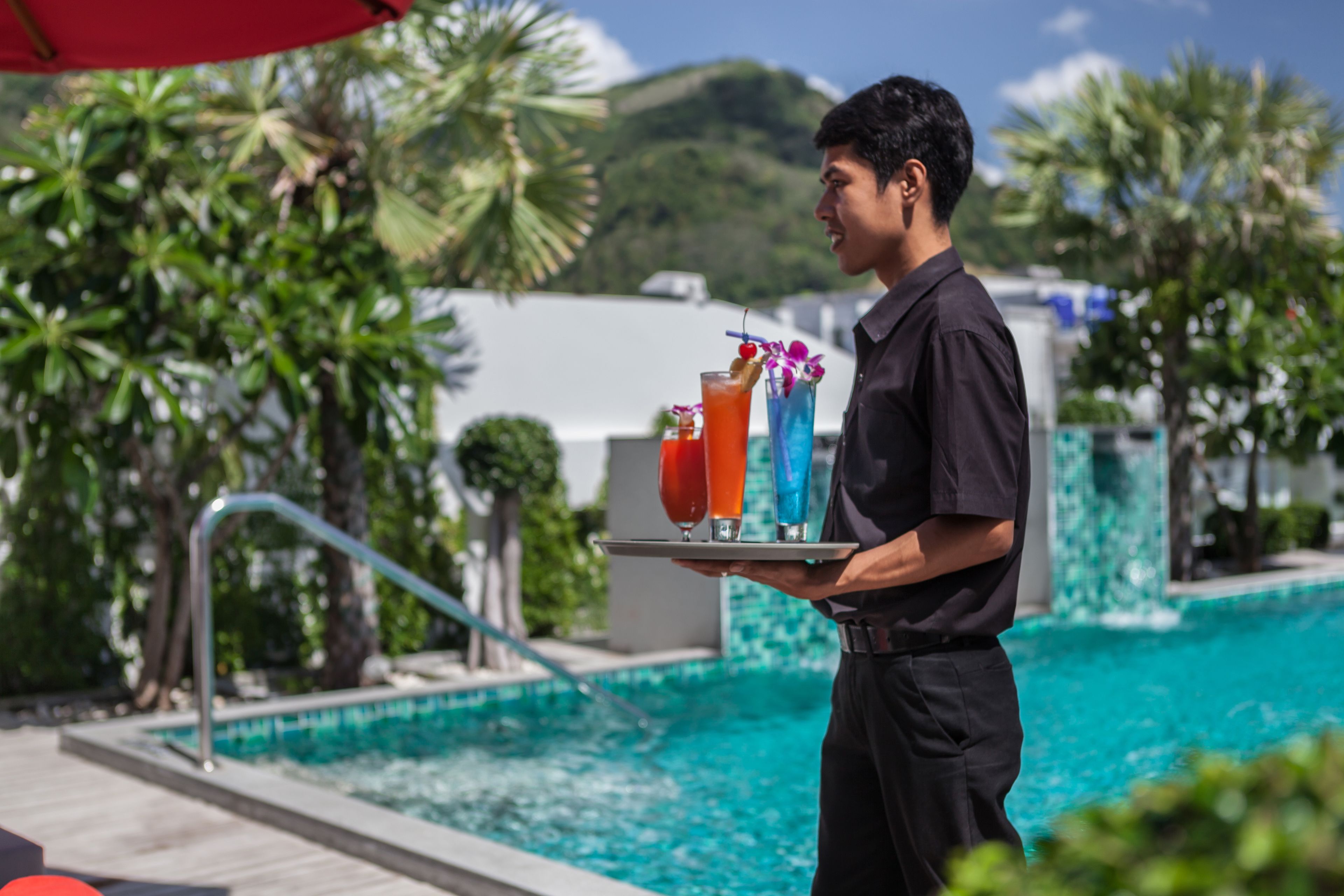 A person holding a tray with drinks near a pool with a clear view of the surrounding greenery and a building in the background.