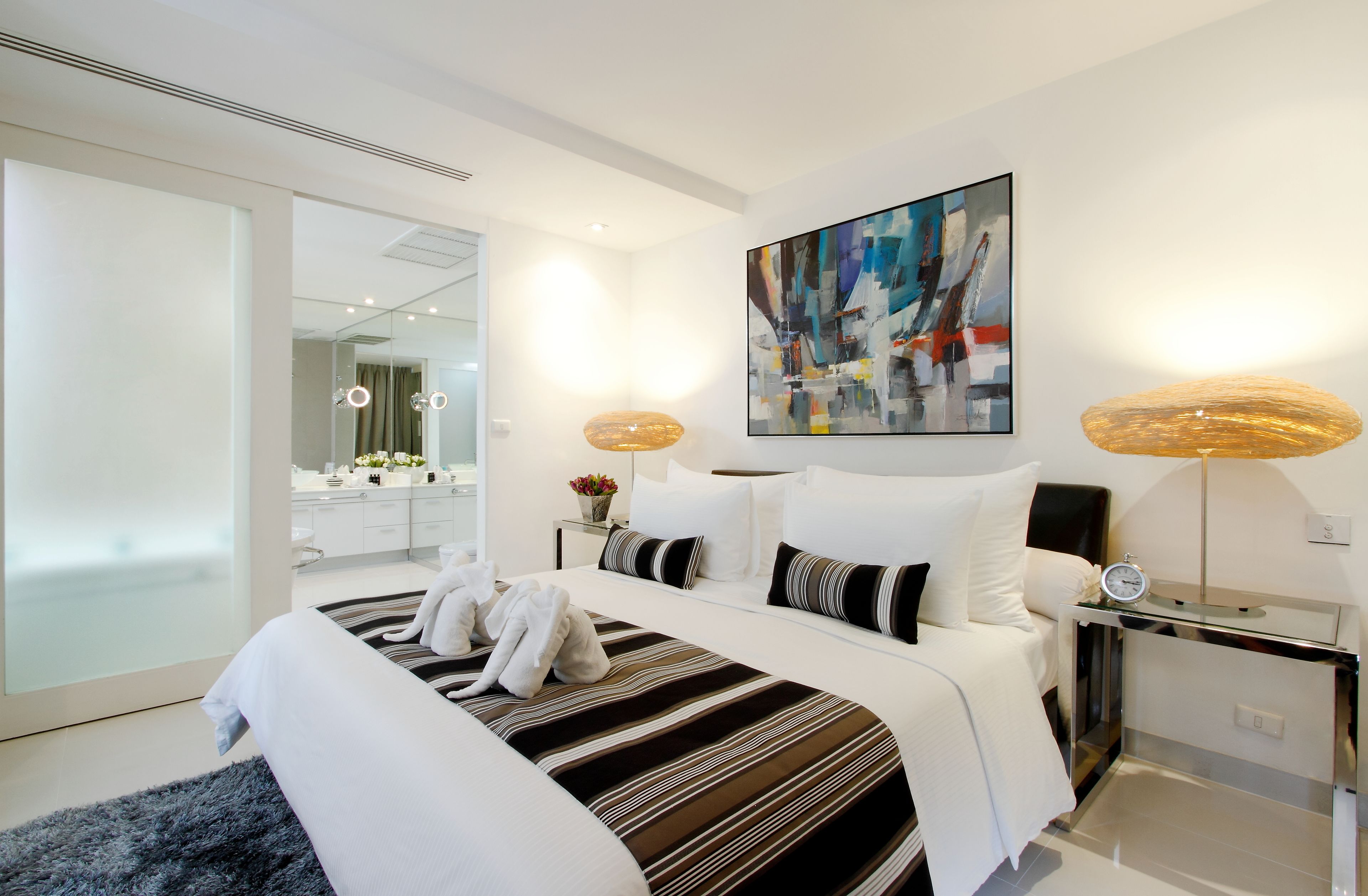 Et moderne hotelværelse med en stor seng, et sengebord med lampe, et billede på væggen og et badeværelse, der kan ses igennem en glasdør.