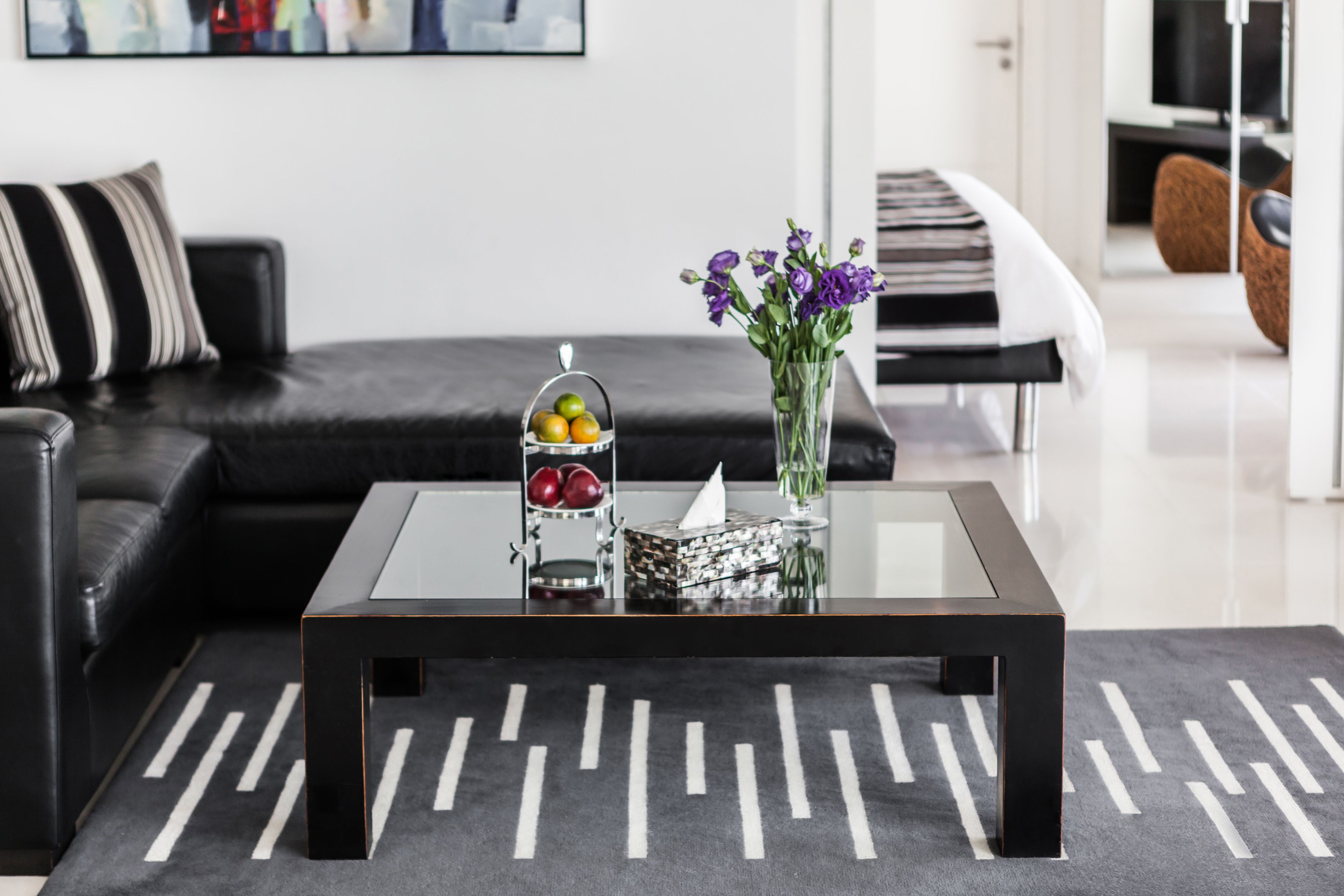Een moderne woonkamer met een glazen salontafel, een zwarte leren bank en een decoratieve vaas met paarse bloemen.