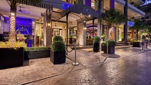 Entrada de hotel com galeria coberta, palmeiras e fachada bem iluminada.
