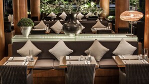 Restaurante com interior moderno, apresentando uma mesa central para refeições com um vaso de folhagem, assentos acolchoados e um espaço de bar com bancada de vidro.