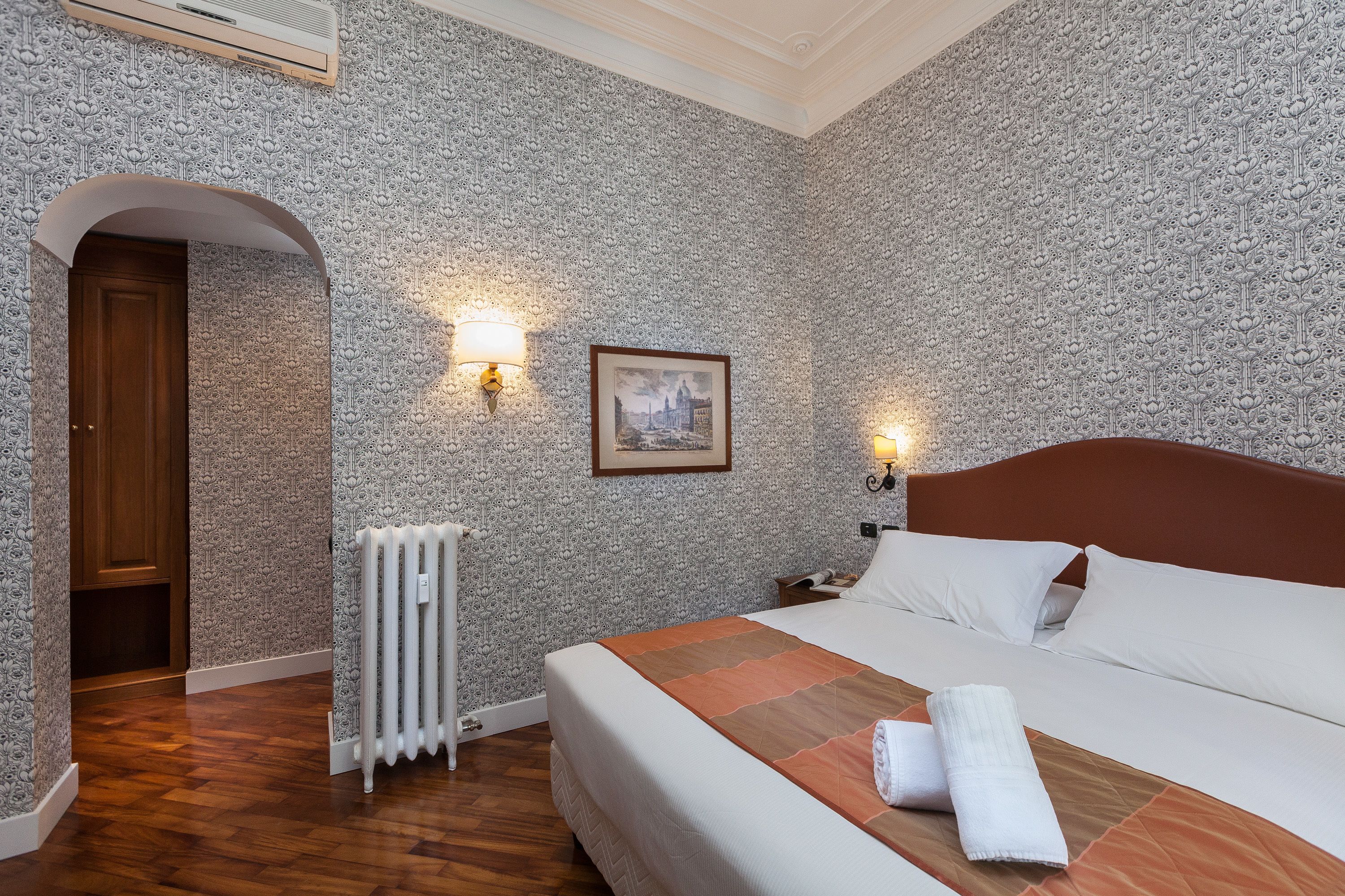 Ein Hotelzimmer mit strukturiertem Wandpapier, einer Holztür, einem Heizkörper, einem gerahmten Bild an der Wand und einem ordentlich bezogenen Bett mit weißen Bettwäsche und braunen Akzenten.