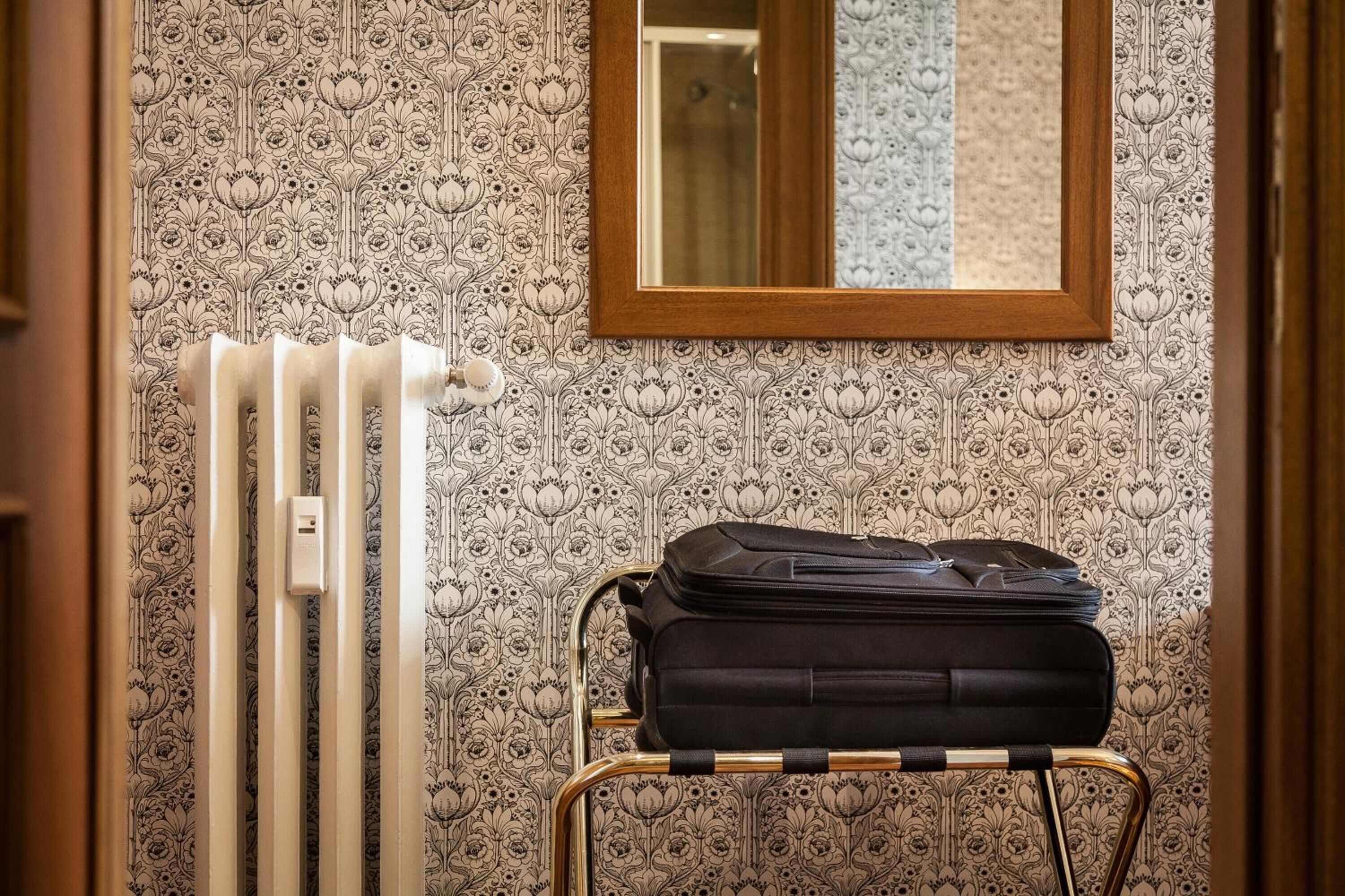Une pièce avec un papier peint à motifs, un radiateur, un miroir et un chariot à bagages.