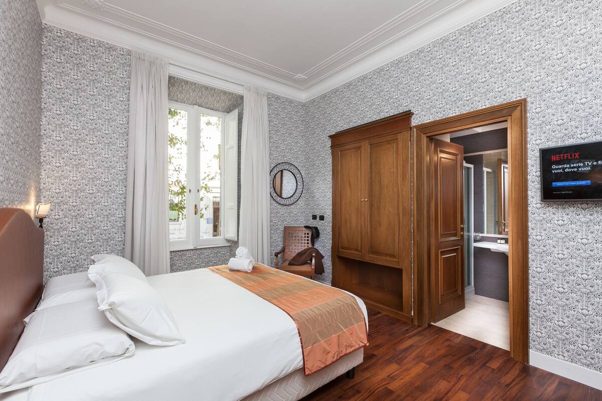 A hotel room with a bed, a wooden wardrobe, a television, and a window with curtains.