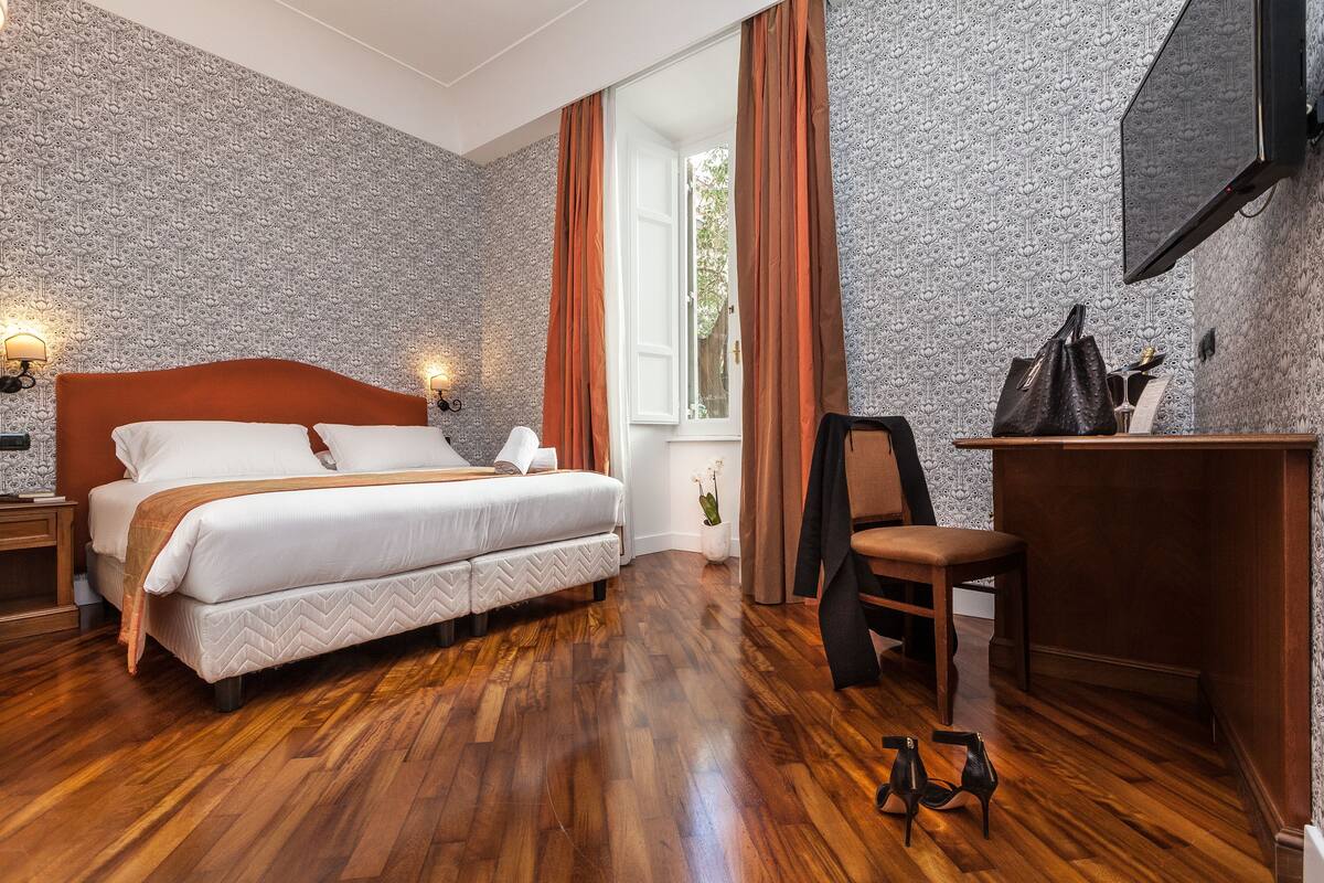 A bedroom with a wooden floor, a bed with a white headboard, a desk with a chair, and a TV mounted on the wall.