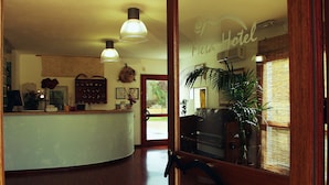 A reception area with a wooden counter, a reception desk, and a large mirror reflecting the interior. There are two pendant lights hanging from the ceiling, and a potted plant is visible in the reflection. - Métà hotel (Santa Teresa di Gallura)