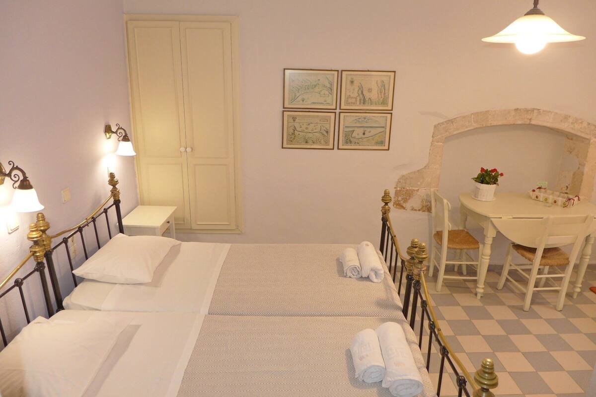 A double bed with white linens, a small bedside table, a chair, a desk with a chair, a wall-mounted lamp, and a checkered floor.
