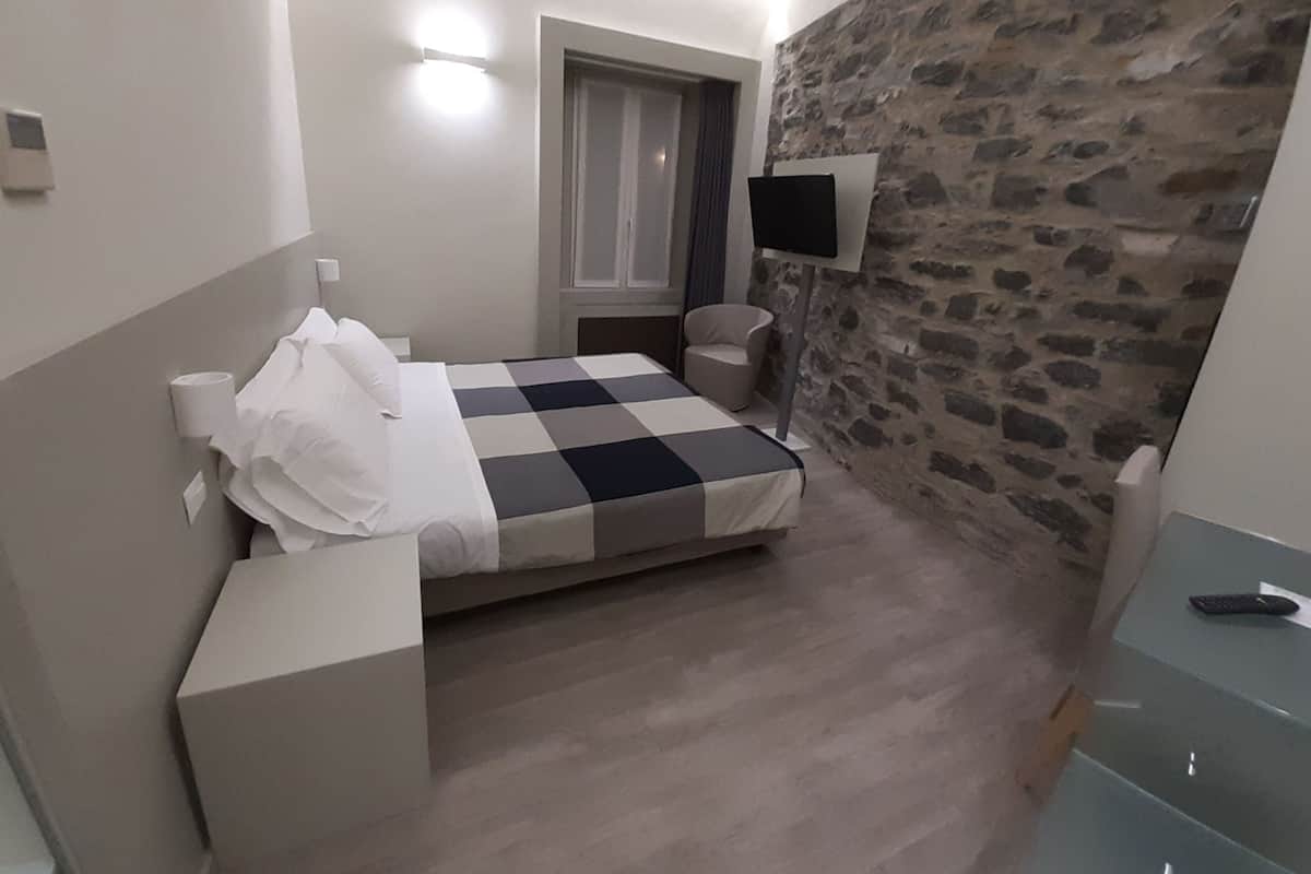 Une chambre avec une couvre-lit à carreaux, une table de chevet, une chaise, un téléviseur fixé au mur en pierre et une fenêtre avec des rideaux.