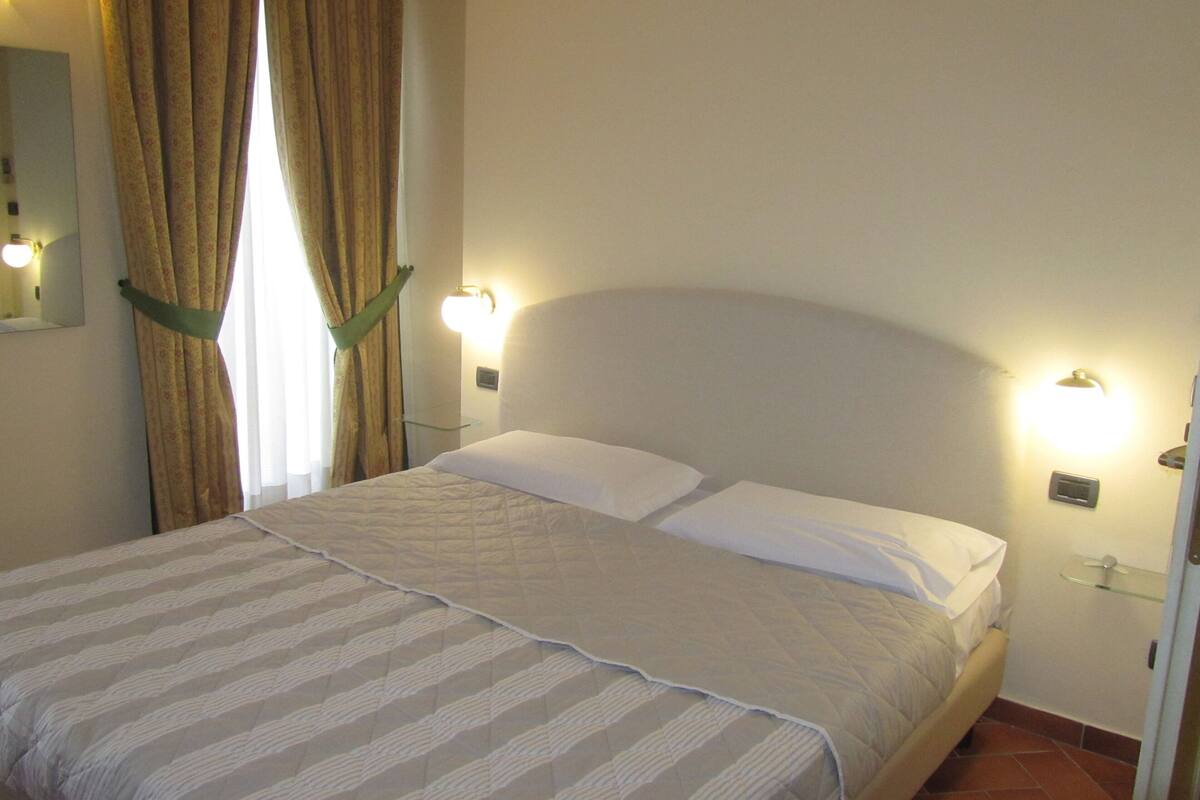 Un lit bien fait, recouvert d’une couvre-lit à rayures, avec deux lampes de chevet et une fenêtre avec des rideaux.