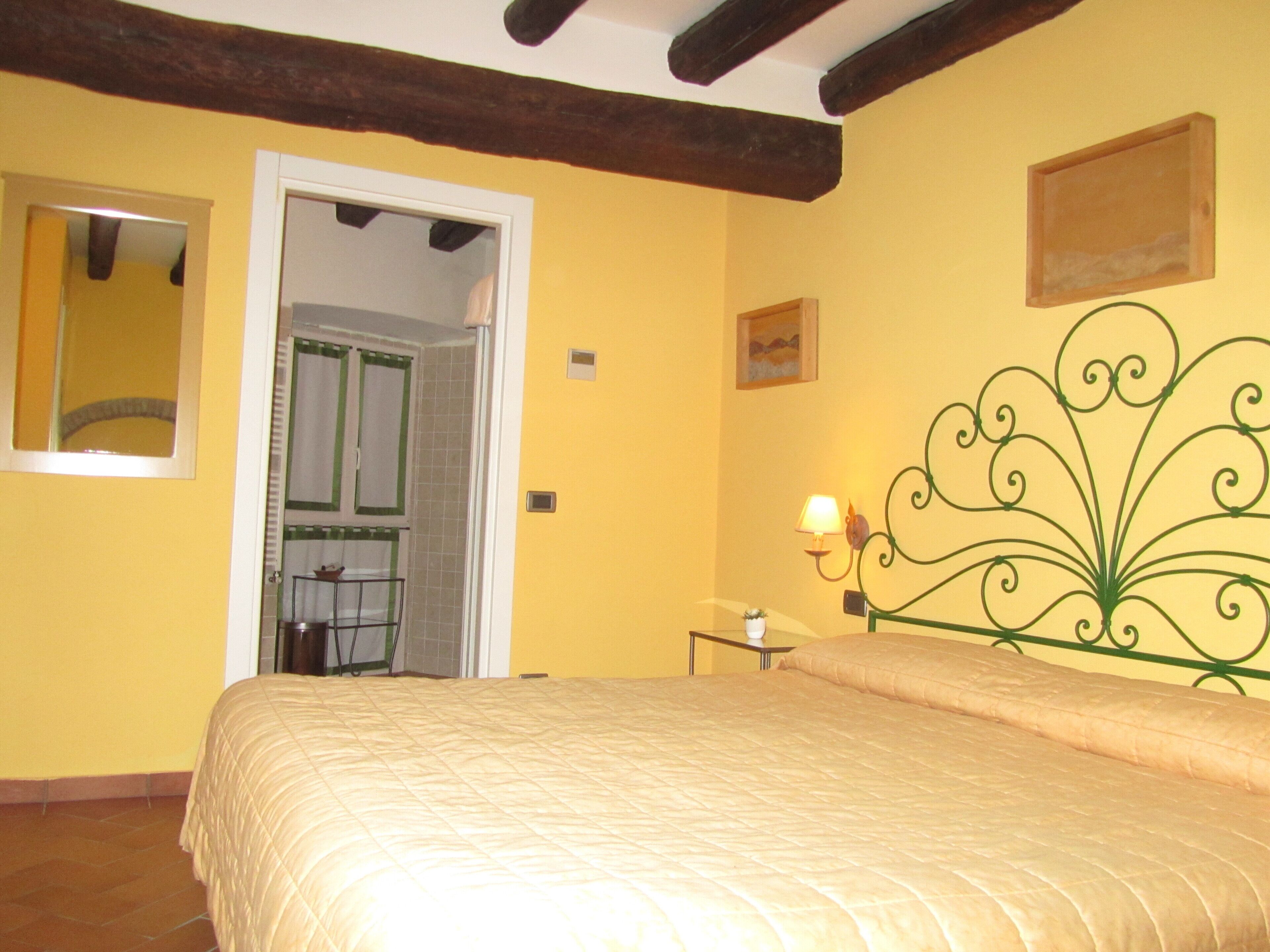 Un dormitorio con una cama, una puerta que da al baño, una lámpara fijada a la pared y una mesita con una planta.